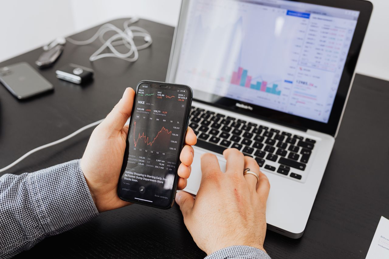 Una persona esamina i dati di borsa con uno smartphone e un laptop, al chiuso.